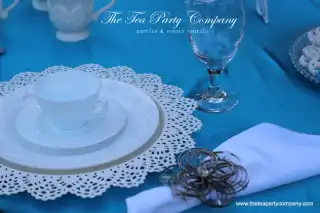 white laced charger with milk glass teacup, saucer and dinner plate  with a silver flower motif napkin holder on top of a blue tablecloth